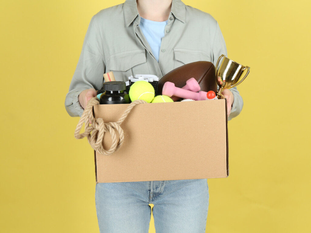 スポーツ用品やトロフィーなどが入った段ボール箱を持つ人